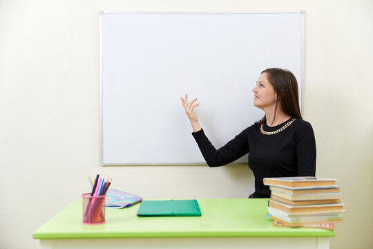 Young teacher sitting at a table near empty white board in classroom
