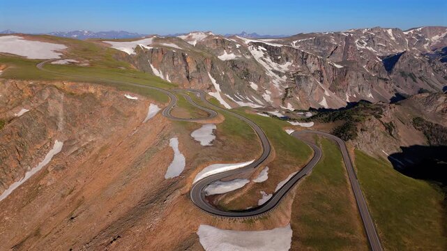 Aerial of Beartooth Highway in Montana USA showing dramatic serpentine mountain road with switchbacks cutting through alpine terrain with snow patches and rugged peaks under clear blue sky.