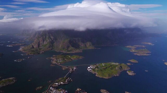 Aerial over Lofoten, Henningsvaer, Norway shows rocky islets, coastal houses, bridges, a small football pitch, and a steep mountain with a layered cap cloud during a gentle pan.