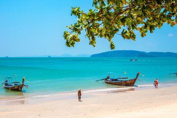 Obraz premium View of a sandy beach with longtail boats and turquoise water through tree branches. tropical landscape, summer vacation, Krabi marine tourism.Krabi, Thailand, March 20, 2024