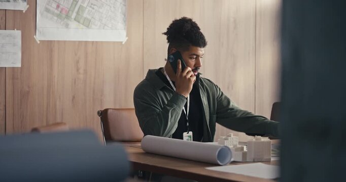 A man sits at a table in an office and speaks on the phone while checking his watch and reviewing plans