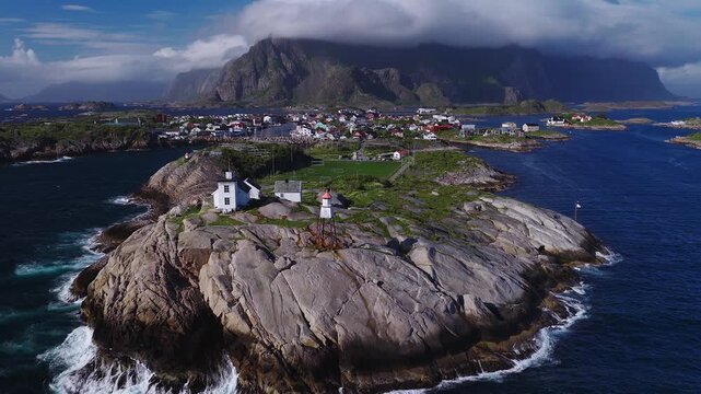 Aerial view of a rocky islet lighthouse and keeper's house as waves hit cliffs, with a compact village and Henningsvaer Stadion in Lofoten, Norway. Mountains under bright summer daylight.