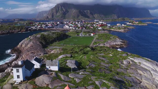 Aerial summer view in Lofoten, Norway shows Henningsvaer Stadion on a rocky islet, red and white houses, fish drying racks, a white lighthouse, harbor, cars, and people.