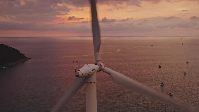 Wind turbine generating electricity with spinning blades while sailboats anchor in the tranquil Andaman Sea along Phuket island, capturing a vibrant sunset sky and symbolizing renewable power