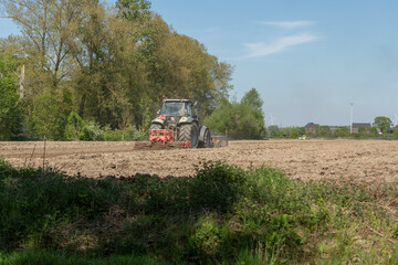 Fototapeta premium Diepenbeek, Belgium. Tractor cultivating large agricultural field with wind turbines in background during spring soil preparation