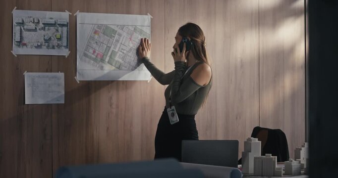A woman stands in an office, talking on the phone and looking over project plans on the wall