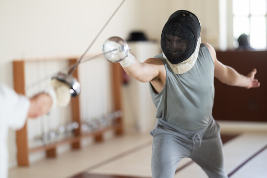 Male fencer wears protective mask, practicing epee techniques with partner in sports hall