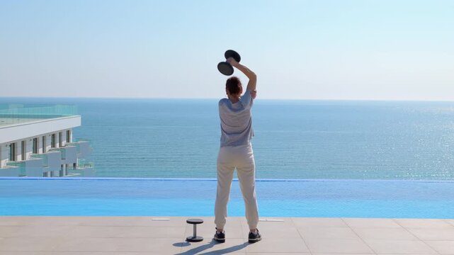 Woman doing a triceps exercise by the pool. Sporty woman performing overhead dumbbell triceps extensions during an outdoor workout on a rooftop with an infinity pool and a beautiful ocean view