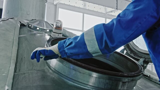 Male brewery worker in a blue uniform opening the large metal hatch of a fermentation tank. Industrial equipment at a modern beer factory during the brewing production process