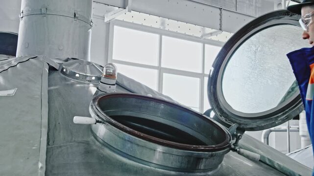 Male brewer in a uniform opening a large stainless steel mash tun hatch to inspect the brewing process inside the modern industrial factory, ensuring quality control of the beverage