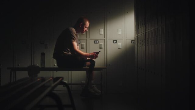 Athlete with Limb Loss Sits on Bench in Dressing Room, Typing Message Trainer on Smartphone. Role of Communication in Professional Sports Recovery. Sports Mentorship and Inclusive Fitness Technology.