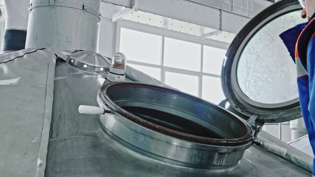 Brewery worker in a uniform opening the hatch of a large stainless steel mash tun tank for inspection. Industrial equipment inside a modern beer production facility during the brewing process