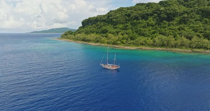 Sailboat yacht anchoring in clear turquoise water near a lush green tropical island coastline, remote travel and adventure in peaceful ocean. Explore Torres islands, Vanuatu. Aerial view drone shot