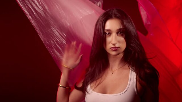 Fashion, beauty, and art concept with a woman posing in a studio, illuminated by red lighting. A model with dark hair holds a translucent plastic sheet, creating an abstract and creative portrait. The