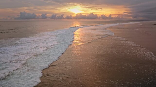 Vibrant orange light from a tropical sunset reflecting on the calm ocean surface and breaking waves along the sandy coastline of Bukit Peninsula, Bali island. Natural landscape. Aerial drone flight