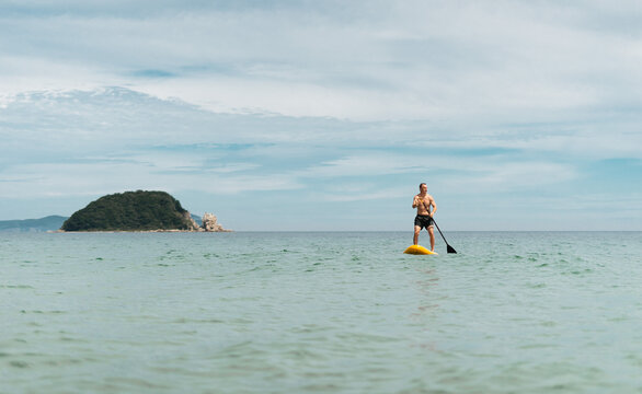 Athletic man enjoying stand up paddle on ocean with scenic island view. Sunny day, fitness activity and leisure travel. Concept of freedom, sport lifestyle and tropical rest. Copy space