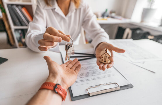 Closeup of woman offering keys and house model during property transaction. Real estate concept illustrating trust, financial planning and achievement of homeownership goals.