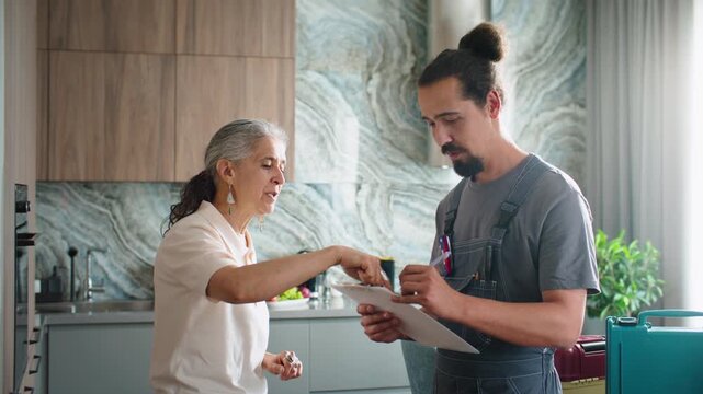 Senior female homeowner explaining her repair needs to young handyman writing down service order details on clipboard in modern kitchen