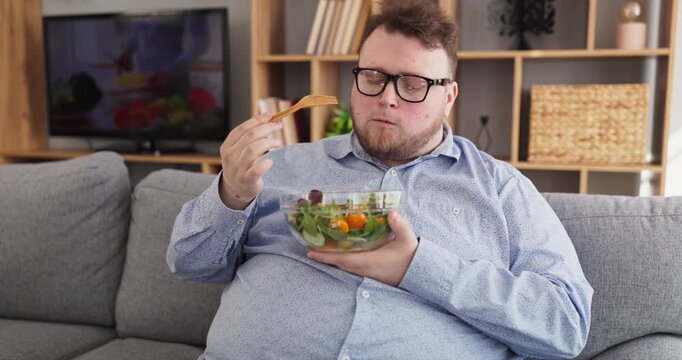 Overweight man eating salad at home sofa. Wearing glasses, he watches tv and maintains a healthy diet. Cozy living room setting supports wellness marketing. Healthy lifestyle change concept.