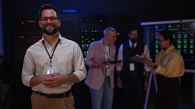 Confident it specialist posing in server room with colleagues working on data analysis in the background