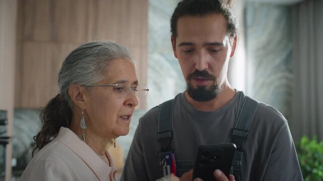 Professional handyman in overalls and senior female client discussing repair estimate on smartphone screen during home maintenance service visit