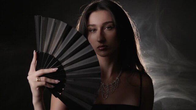 Fashion and beauty portrait of a woman holding a black fan, elegance and glamour. The studio shot features dramatic lighting and a smoke effect, creating a moody and sensual atmosphere. Commercial, ed