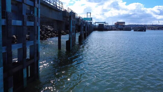 Drone footage following a harbor jetty as seagulls soar above calm waters near industrial docks and breakwaters in Matane, Quebec, Canada.