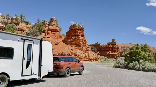 A burgundy SUV with a travel trailer RV parked at the entrance of Dixie National Forest. Majestic red rocks and clear blue sky on a sunny day. American road trip and camper lifestyle concept.