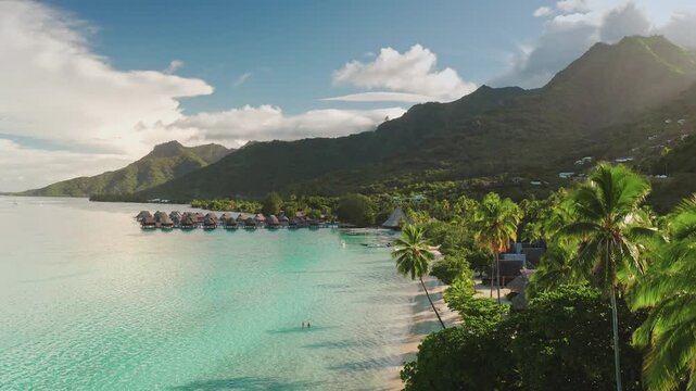 Moorea island showcasing overwater bungalows lining a pristine white sand beach, with turquoise ocean waters, lush green mountains, and swaying palm trees under a bright blue sky. Tropical paradise