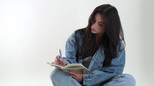 Creative writing and education concept with a young woman student diligently writing in a notebook with a pen. The model is casually dressed in denim, sitting on the floor, and isolated on a clean whi