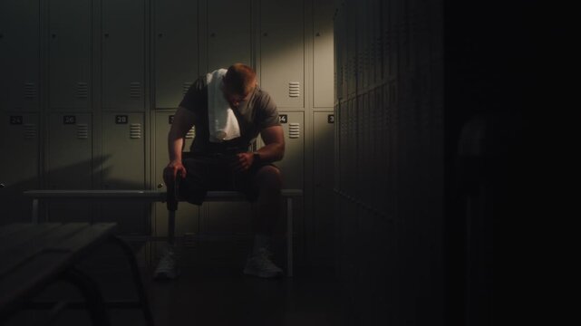 Man Heavily Sits Down on Bench, Leaning Back Against Metal Lockers For Support. Male Taking Deep, Labored Breaths Recover While Hydrating From Water Bottle. Grit, Determination of Resilient Athlete.