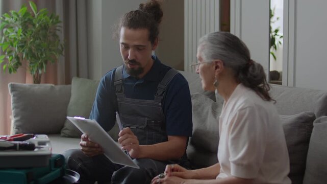 Young handyman in work overalls and senior female client speaking and signing repair contract while sitting in living room at home