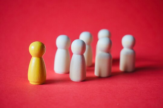 Leadership and career individuality concept with a distinct yellow painted wooden figure standing out from a crowd of white figures on a red background.