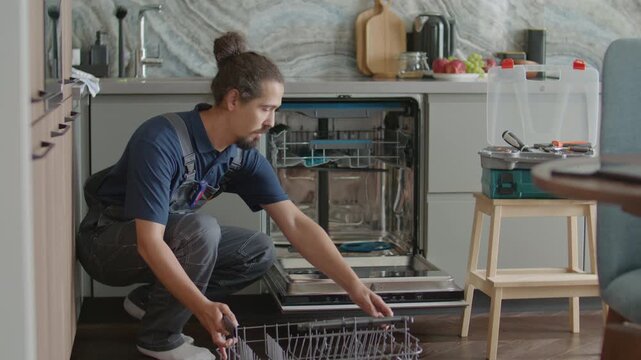 Young professional handyman crouching down in front of open dishwasher, inspecting internal parts during home service visit, his toolbox placed on stool nearby in kitchen