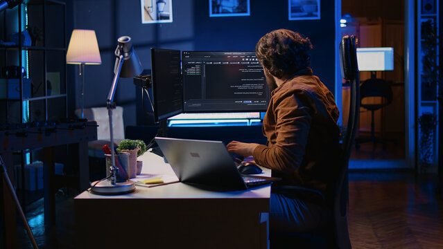 Software programmer putting headphones on while developing code, making time pass faster during remote job shift. IT professional doing multitasking in apartment office while listening music.