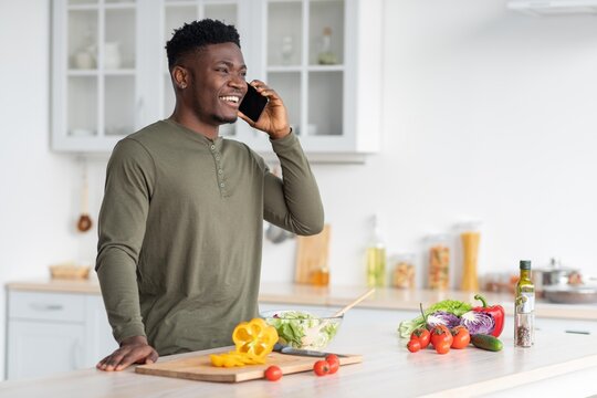 Smiling Black Man In Casual Attire Talking With Smartphone While Preparing Healthy Salad Ingredients In Modern Kitchen. Engaged And Happy Young Adult Enjoying Cooking Session At Home.