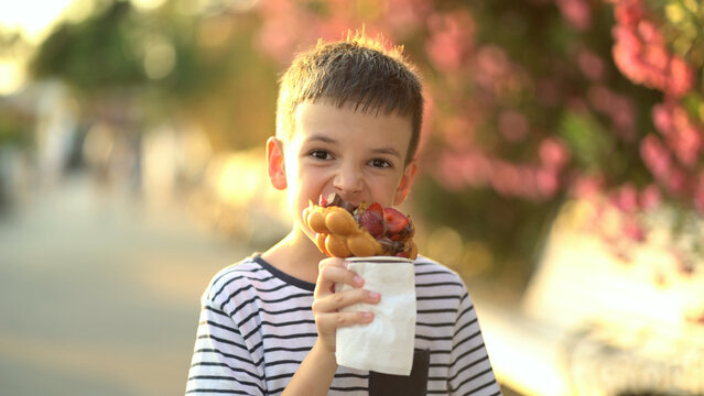 A cute young boy happily eats a bubble waffle topped with chocolate sauce and fresh strawberries outdoors at golden hour.