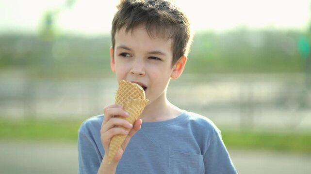 Cute little boy eats green ice cream cone outdoors making funny playful face with squinted eye biting waffle cone on sunny summer day.