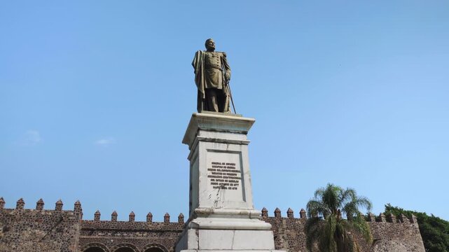 General Carlos Pacheco monument viewed from a low angle in Cuernavaca with the Regional Museum of the Peoples behind it. The scene highlights history and architecture under bright daylight.