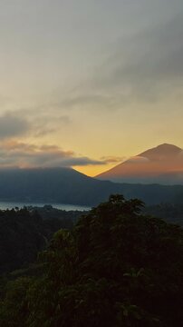 Vertical video. Timelapse Sunrise View of Gunung Batur and Gunung Abang With Fast Moving Clouds Above Lake and Forested Highlands Under Golden Morning Light