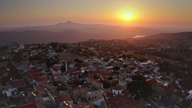 Pano Lefkara village nestled in Troodos Mountains at sunset, traditional stone houses and red roofed church with bell tower glowing in warm golden light over peaceful valley. Aerial view drone flight