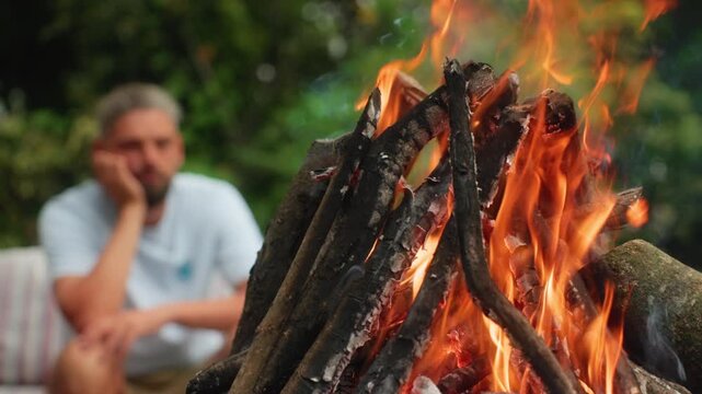 Close-up of crackling campfire with orange flames and burning wood logs. Blurred man in white shirt rests chin on hand while sitting outdoors in garden setting with green foliage.