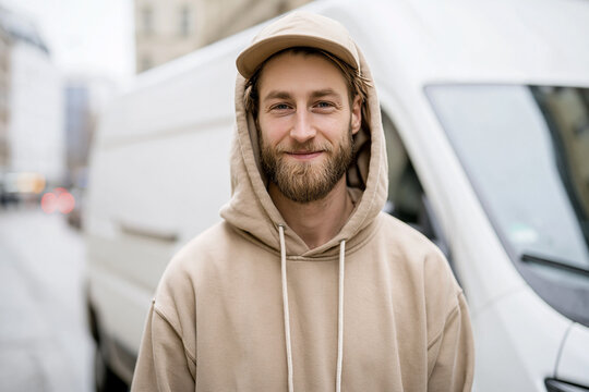  Portrait d'un jeune livreur souriant portant une casquette et un sweat &agrave; capuche devant une camionnette de livraison blanche en ville.