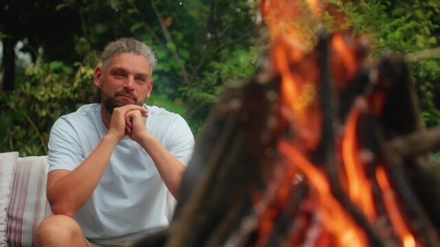 Thoughtful bearded man with gray curly hair relaxes outdoors beside a crackling bonfire with bright orange flames. Lush green foliage fills background as he sits on a cushioned seat in contemplation.