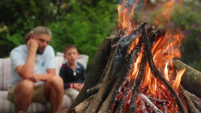 Father and son relax by campfire in lush green backyard garden. Burning wood logs create bright orange flames and glowing embers as family enjoys peaceful outdoor bonding time together on summer day.