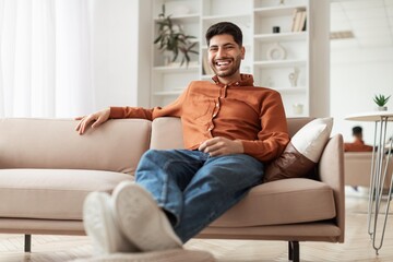 Obraz premium Smiling Young Caucasian Man Relaxing On Couch In Cozy Modern Living Room, Enjoying Comfort And Leisure While Sitting Casually, Inviting Atmosphere Of Home. Happiness Concept.