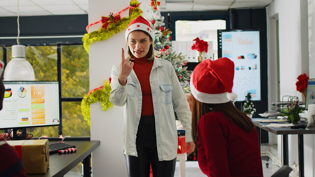 Employees wearing Santa Claus hat enjoying Christmas spirit mood playing pretending acting game during break, coworkers having fun trying to guess actions during pantomime in Christmas adorn office.