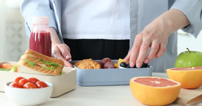 Woman putting bowl of fresh blueberries into lunch box and showing it at light wooden table indoors, closeup