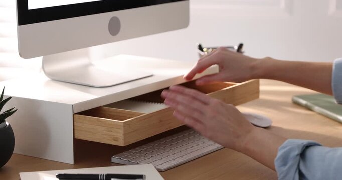 Woman taking glasses out of under monitor riser at wooden desk indoors, closeup