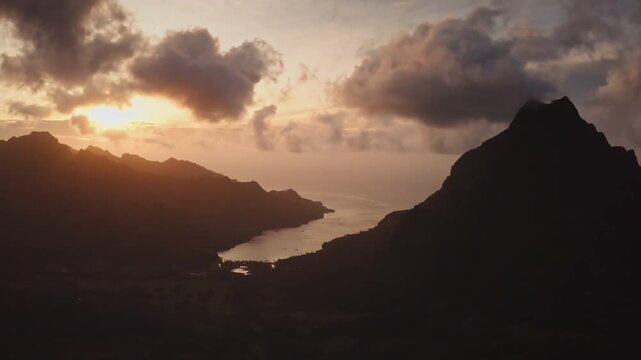 Breathtaking aerial view capturing the striking silhouettes of the mountains on Moorea, French Polynesia, as the sun dips below the horizon, bathing the sky and tranquil bay in warm, golden light